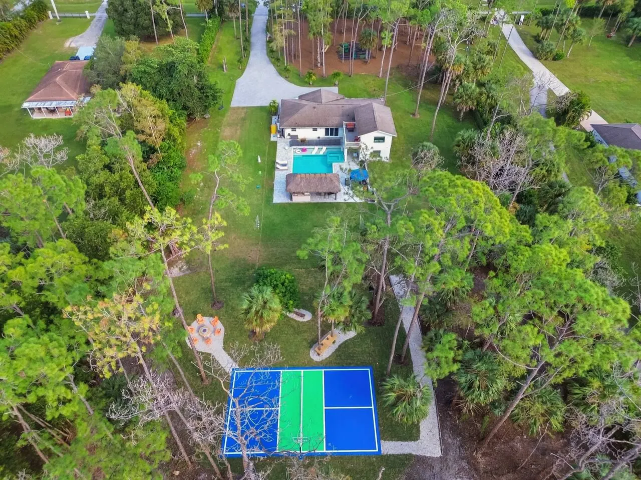 Overhead drone view of the property: house and pool at the top, open lawn, fire pit and sports court among tall pines.